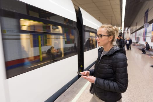 Young casual woman with a cell phone in her hand waiting on the platform of a metro station for metro to arrive. Public transport.