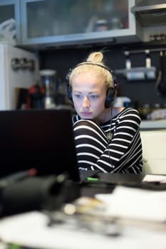 Woman in her casual home clothing working and studying remotely from her small flat late at night. Home kitchen in background. Great flexibility of web-based courses and study programmes.