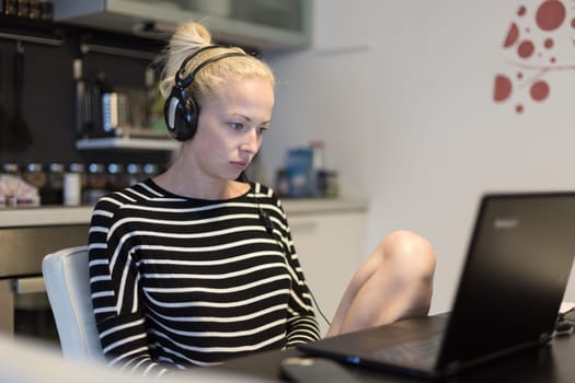 Woman in her casual home clothing working and studying remotely from her small flat late at night. Home kitchen in background. Great flexibility of web-based courses and study programmes.