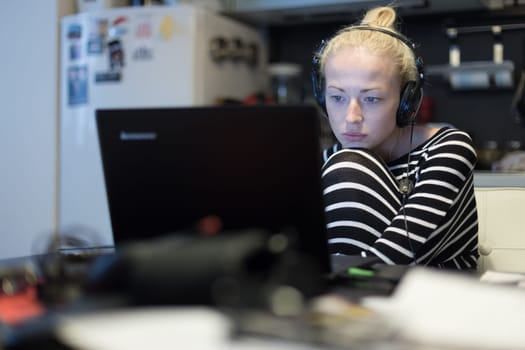 Woman in her casual home clothing working and studying remotely from her small flat late at night. Home kitchen in background. Great flexibility of web-based courses and study programmes.