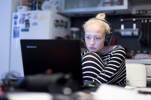 Woman in her casual home clothing working and studying remotely from her small flat late at night. Home kitchen in background. Great flexibility of web-based courses and study programmes.