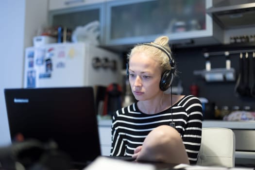 Woman in her casual home clothing working and studying remotely from her small flat late at night. Home kitchen in background. Great flexibility of web-based courses and study programmes.