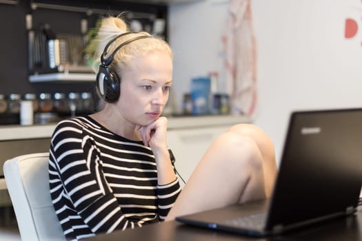 Woman in her casual home clothing working and studying remotely from her small flat late at night. Home kitchen in background. Great flexibility of web-based courses and study programmes.