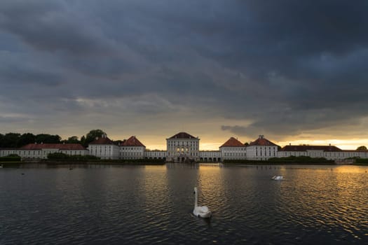 Dramatic scenery of Nymphenburg palace in Munich Germany. Sunset after the sorm. White swans and duks swimming in pond in front of the palace.