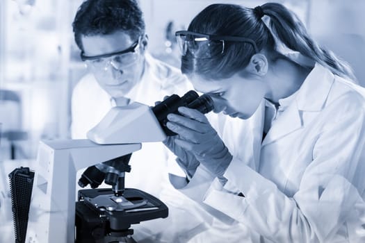 Life scientists researching in laboratory. Attractive female young scientist and her doctoral supervisor microscoping in their working environment. Healthcare and biotechnology. Greyscale blue toned.