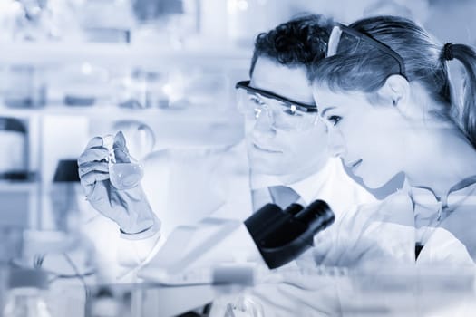 Chemical laboratory. Attractive young PhD student and her post doctoral supervisor scientist observing the blue indicator color shift after the solution destillation. Greyscale blue toned image.