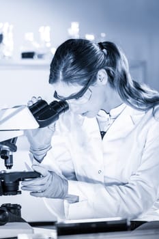 Life scientists researching in laboratory. Attractive female young scientist microscoping in their working environment. Healthcare and biotechnology. Greyscale blue toned image.