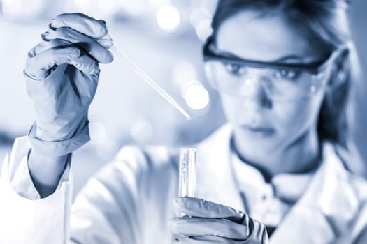 Life scientists researching in laboratory. Focused female science professional pipetting solution into glass cuvette. Lens focus on pipette. Healthcare and biotechnology. Greyscale blue toned image.