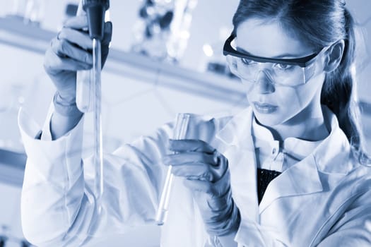 Life scientists researching in laboratory. Focused female life science professional pipetting solution into glass cuvette. Healthcare and biotechnology. Greyscale blue toned image.