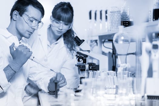 Young male researcher pipetting red liquid in the glass tube in the laboratory. Female asistant science student watching and learning the protocol.