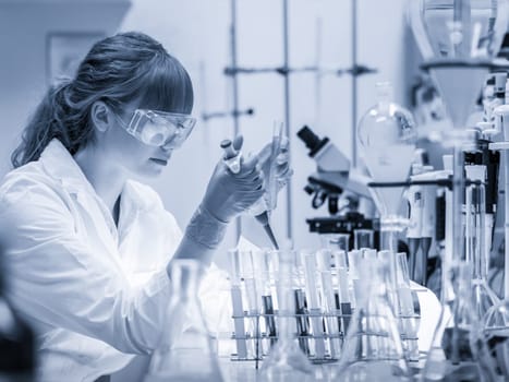 Life scientists researching in laboratory. Focused female science professional pipetting solution into glass cuvette. Lens focus on pipette. Healthcare and biotechnology. Greyscale blue toned image.