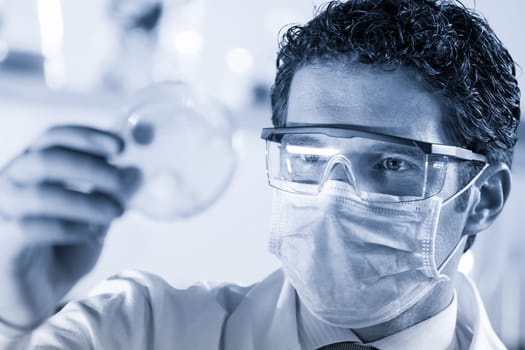 Mask and goggles protected life science researcher observing potentially infectious cells in petri dish. Focus on scientist's eye. Healthcare and biotechnology concept. Greyscale blue toned image.