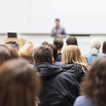 Male speaker giving presentation in lecture hall at university workshop. Audience in conference hall. Rear view of unrecognized participant in audience. Scientific conference event.