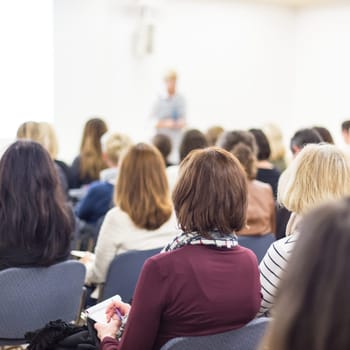 Business and entrepreneurship symposium. Female speaker giving a talk at business meeting. Audience in conference hall. Rear view of unrecognized participant in audience. Copy space on whitescreen.