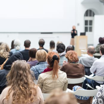 Business and entrepreneurship symposium. Female speaker giving a talk at business meeting. Audience in conference hall. Rear view of unrecognized participant in audience. Copy space on whitescreen.
