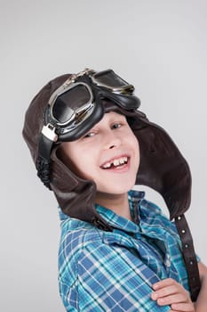 Against a grey background,the exposed upper body view of a ten-year-old boy in a blue-checkered short-sleeved shirt and brown leather cap with classic motorcycle glasses smiling joyfully into the camera.