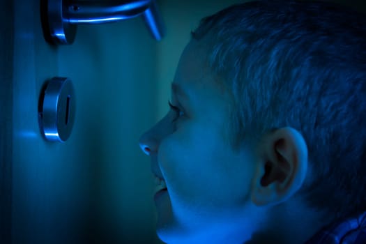 Boy in the dark room looks through a keyhole in the glow of the flashlight.