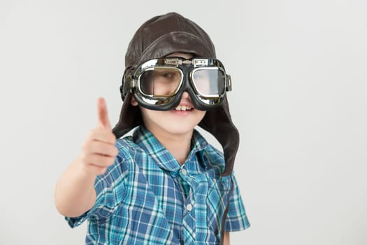 Against a grey background,the exposed upper body view of a ten-year-old boy in a blue-checkered short-sleeved shirt and brown leather cap with classic motorcycle glasses smiling joyfully into the camera. Thumbs up.