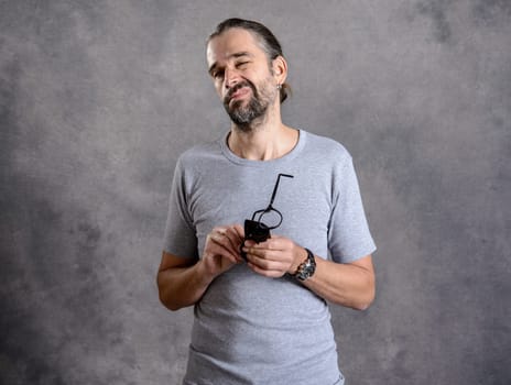 young bearded man ceaning his glasses in front of gray background