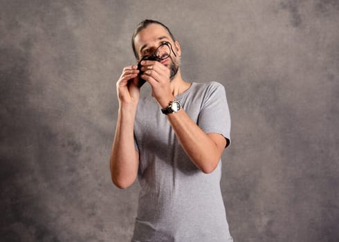 young bearded man ceaning his glasses in front of gray background