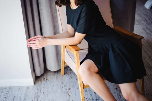 Young attractive woman with long legs in black elegant dress, sits in chair near window in interior of room.