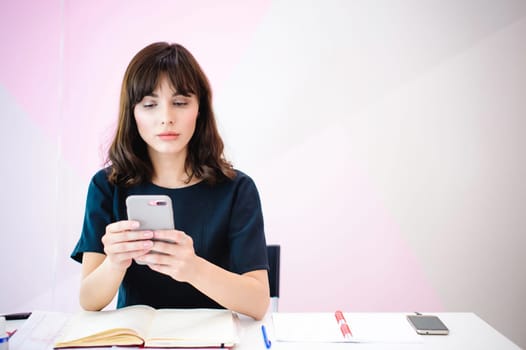Portrait of a beautiful business woman making notes in a smartphone. Holding a cell phone while sitting in a workplace in a pink office