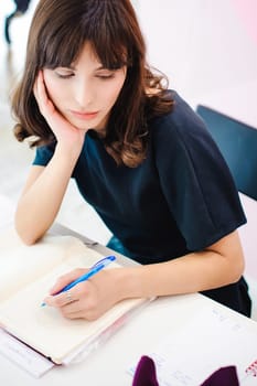 Portrait of a beautiful business woman making notes in a notebook. Holds a pen, sitting in the workplace in a pink office