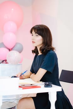 Portrait of a beautiful business woman making notes in a notebook. Holds a pen, sitting in the workplace in a pink office