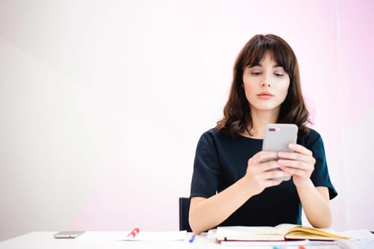 Portrait of a beautiful business woman making notes in a smartphone. Holding a cell phone while sitting in a workplace in a pink office