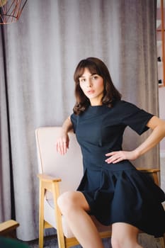 Young attractive woman with long legs in black elegant dress, sits in chair near window in interior of room.