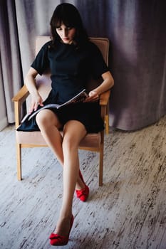 Young attractive woman with long legs in black elegant dress, sits in chair near window in interior of room.