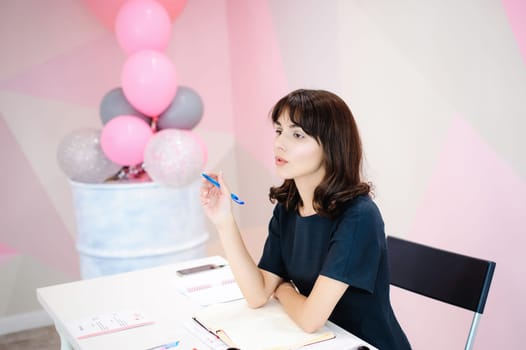 Portrait of a beautiful business woman making notes in a notebook. Holds a pen, sitting in the workplace in a pink office