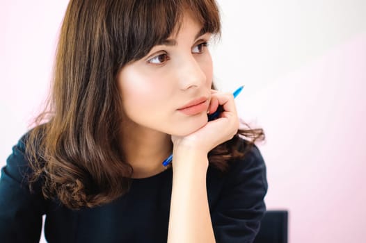 Portrait of a beautiful business woman making notes in a notebook. Holds a pen, sitting in the workplace in a pink office