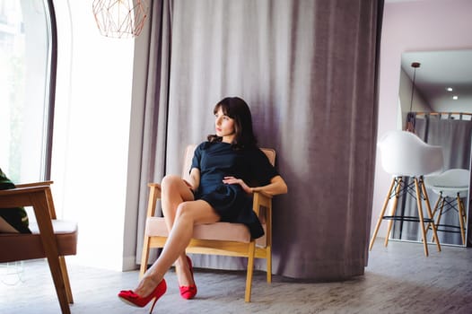 Young attractive woman with long legs in black elegant dress, sits in chair near window in interior of room. Seductive shoes of red light, on beautiful legs