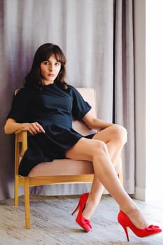 Young attractive woman with long legs in black elegant dress, sits in chair near window in interior of room. Seductive shoes of red light, on beautiful legs