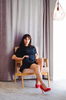 Young attractive woman with long legs in black elegant dress, sits in chair near window in interior of room. Seductive shoes of red light, on beautiful legs