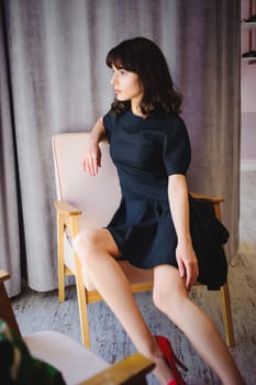Young attractive woman with long legs in black elegant dress, sits in chair near window in interior of room.