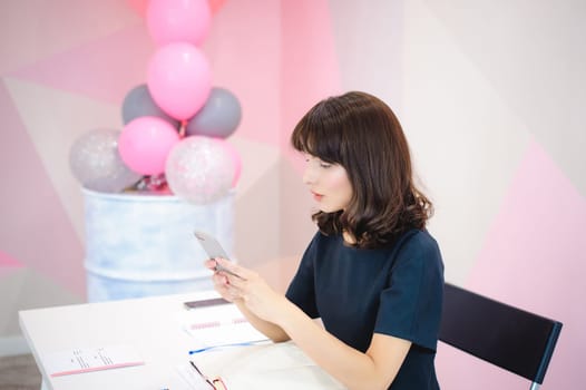 Portrait of a beautiful business woman making notes in a smartphone. Holding a cell phone while sitting in a workplace in a pink office