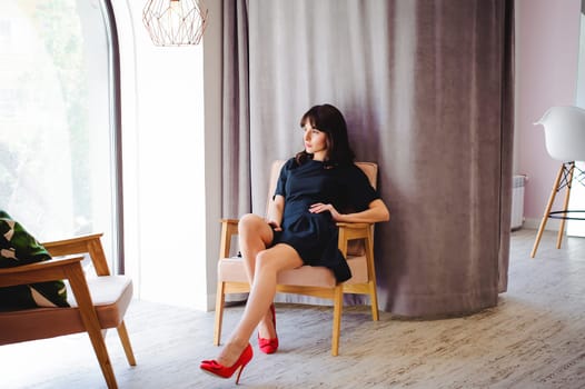 Young attractive woman with long legs in black elegant dress, sits in chair near window in interior of room. Seductive shoes of red light, on beautiful legs