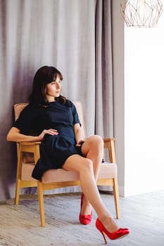 Young attractive woman with long legs in black elegant dress, sits in chair near window in interior of room. Seductive shoes of red light, on beautiful legs
