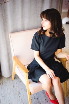 Young attractive woman with long legs in black elegant dress, sits in chair near window in interior of room. Seductive shoes of red light, on beautiful legs