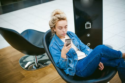 young beautiful woman in jeans clothes in business space of shopping center. portrait of a girl with freckles on her face, stylish girl sits in an armchair with a cell phone in wi-fi zone