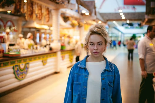 young beautiful woman in jeans clothes in business space of shopping center. portrait of a girl with freckles on her face, stylish girl standing near show-windows of fair in shopping center