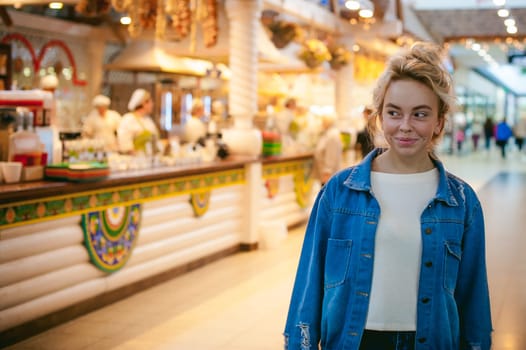 young beautiful woman in jeans clothes in business space of shopping center. portrait of a girl with freckles on her face, stylish girl standing near show-windows of fair in shopping center