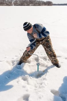 Winter fishing on the river. Young man drill a hole in the ice.