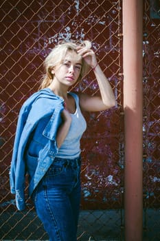 young woman in jeans clothes outdoors. portrait of a girl with freckles on her face, stylish girl on a background of a fence of a grid, on a sunny summer day. Holds a denim jacket in his hand