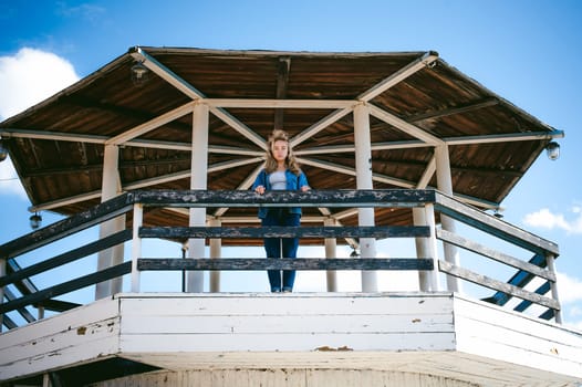 young beautiful woman in jeans clothes outdoors. portrait of a girl with freckles on her face, stylish girl on sea beach rescue tower, on a sunny summer autumn day.