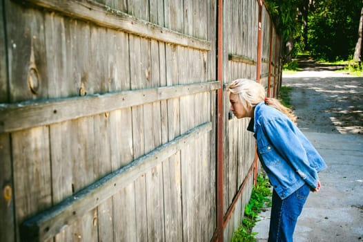 young woman in jeans clothes outdoors. a portrait of a girl with freckles on her face, peeps through a hole in a wooden fence, on a sunny summer day.