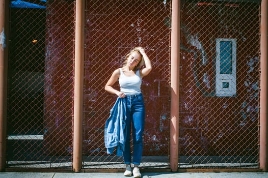 young woman in jeans clothes outdoors. portrait of a girl with freckles on her face, stylish girl on a background of a fence of a grid, on a sunny summer day. Holds a denim jacket in his hand