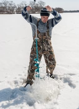 Winter fishing on the river. Young man drill a hole in the ice.